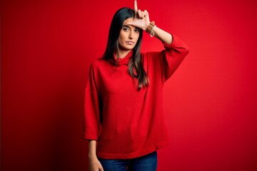 Young beautiful brunette woman wearing casual sweater over isolated red background making fun of people with fingers on forehead doing loser gesture mocking and insulting.