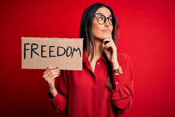 Young beautiful brunette activist woman wearing glasses protesting for freedom holding poster...