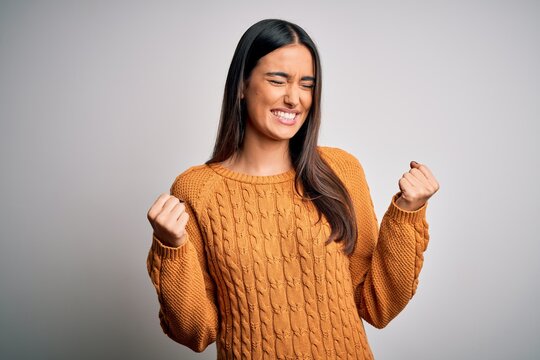 Young beautiful brunette woman wearing casual sweater over isolated white background very happy and excited doing winner gesture with arms raised, smiling and screaming for success. Celebration