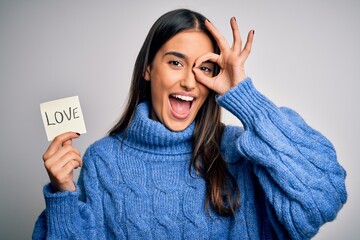 Young beautiful brunette romantic woman holding reminder paper with love message with happy face smiling doing ok sign with hand on eye looking through fingers