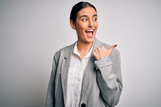 Young beautiful brunette businesswoman wearing jacket standing over white background smiling with happy face looking and pointing to the side with thumb up.