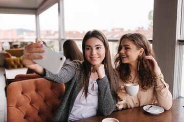Attractive young women making selfie in restairant. Women drin cappuccino on terrace. Two female make photo together