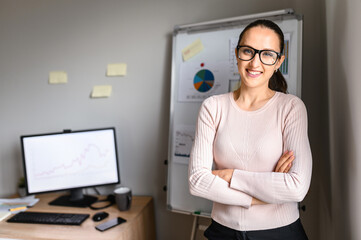 Horizontal middle shot of a female employee with hands crossed around her waist at the office and looks in camera, wearing glasses, flip chart on the background. Consummate employee stands in office.