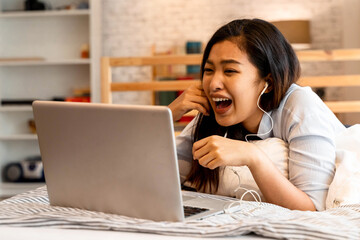 Young smiling Asian woman in casual clothing and lying on bed with laptop computer. Female freelancer working from home on notebook