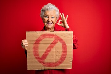 Senior beautiful grey-haired woman holding banner with prohibited signal over red background doing...
