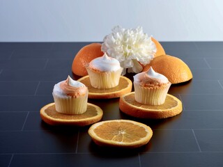 Mini orange creamsicle yellow cupcakes with orange and white frosting sitting on top of fresh cut orange fruit slices with a white artificial flower and orange halves for decoration on black tile.