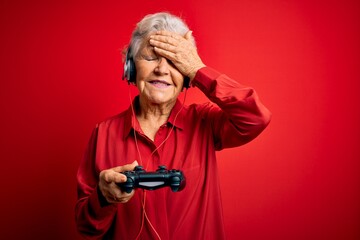 Senior beautiful grey-haired gamer woman playing video game using joystick and headphones stressed with hand on head, shocked with shame and surprise face, angry and frustrated. Fear and upset