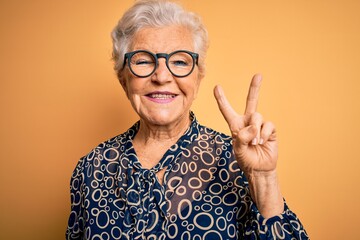 Senior beautiful grey-haired woman wearing casual shirt and glasses over yellow background smiling...