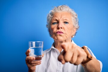 Senior beautiful woman drinking glass of water standing over isolated blue background pointing with finger to the camera and to you, hand sign, positive and confident gesture from the front