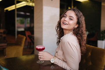 Young woman sitting in restaurant and drink cocktail. Beautiful girl with curly hair smile to camera