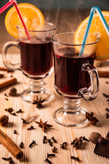 Differential focus. Limpid glasses with hot mulled wine, cloves, fragrant cinnamon sticks,anise flowers stars, nutmegs, lemon and orange on structured natural wooden board, close up front view.