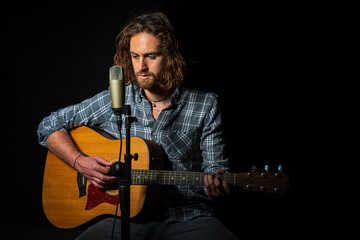 attractive man with long hair with blue eyes sitting next to a microphone and playing acoustic guitar dressed in lumberjack plaid shirt on black background