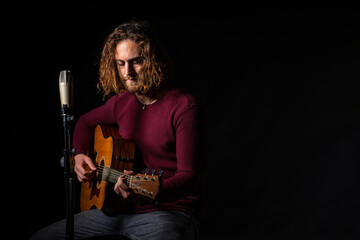 attractive man with long hair with blue eyes sitting next to a microphone and playing acoustic guitar on black background