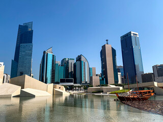 A modern looking cityscape of the Abu-Dhabi with couple of skyscrapers, a modern-looking fountain, and a boat.