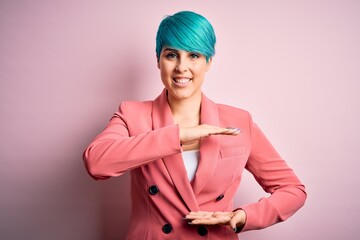 Young beautiful businesswoman with blue fashion hair wearing jacket over pink background gesturing with hands showing big and large size sign, measure symbol. Smiling looking at the camera. Measuring