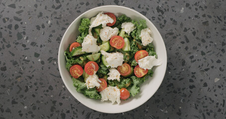 Top view salad with kale, tomatoes cucumbers, and mozzarella in white ceramic bowl on concrete background