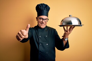 Middle age handsome grey-haired waiter man wearing cooker uniform and hat holding tray approving doing positive gesture with hand, thumbs up smiling and happy for success. Winner gesture.