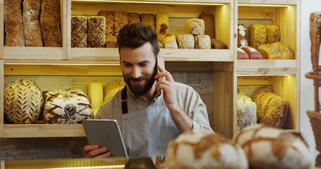 Portrait of happy baker working in bakery shop using digital tablet computer and talking on cell phone
