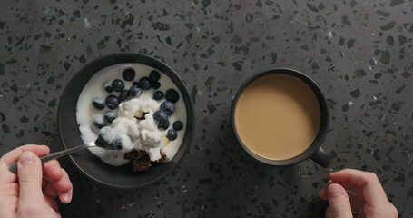 top view man hands eating chocolate granola with almond flakes and blueberry in black bowl and drinking coffee on terrazzo surface