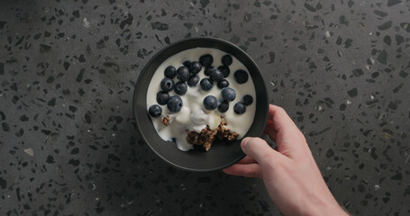 top view man hands put chocolate granola, almond flakes and blueberry in black bowl on terrazzo surface