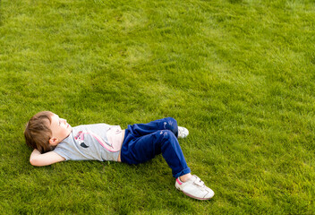 Fototapeta na wymiar a little girl lies on the green grass on her back, hands behind her head, looking up at the sky, dreaming. The child is dressed in a grey t-shirt with a pink Flamingo, blue trousers with a pattern 