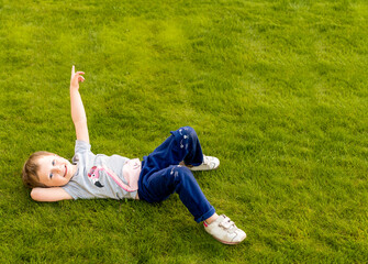 Fototapeta na wymiar a little girl is lying on the green grass on her side, looking at the camera, smiling. The hand is pointing up. The child is dressed in a grey t-shirt with a pink Flamingo, 