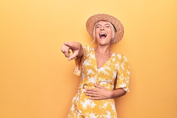 Beautiful blonde woman on vacation wearing summer hat and dress over yellow background laughing at you, pointing finger to the camera with hand over body, shame expression