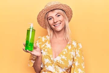 Beautiful blonde woman on vacation wearing summer hat holding bottle with aloe vera cream looking positive and happy standing and smiling with a confident smile showing teeth
