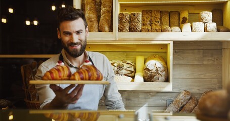 Portrait of the handsome young male baker smiling while posing with croissants on the tray in the shop. Indoors