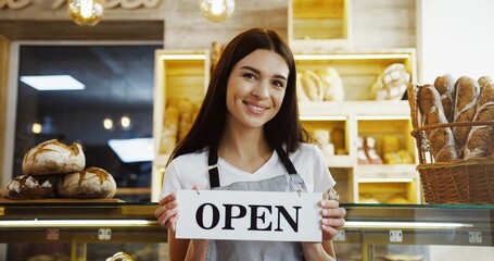 Pretty female baker in glasses scrolling and taping on the tablet computer while standing at the counter in the shop. Indoor