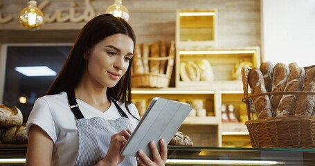 Pretty female baker in glasses scrolling and taping on the tablet computer while standing at the counter in the shop. Indoor