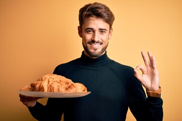 Young man with beard holding plate with croissants standing over isolated yellow background doing ok sign with fingers, excellent symbol