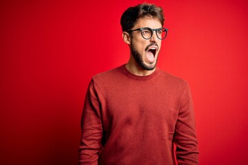 Young handsome man with beard wearing glasses and sweater standing over red background angry and mad screaming frustrated and furious, shouting with anger. Rage and aggressive concept.