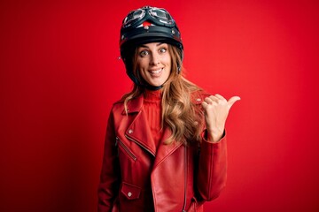Young beautiful brunette motrocyclist woman wearing moto helmet over red background smiling with happy face looking and pointing to the side with thumb up.