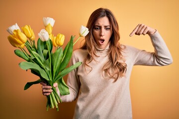 Young beautiful brunette woman holding bouquet of yellow tulips over isolated background Pointing down with fingers showing advertisement, surprised face and open mouth