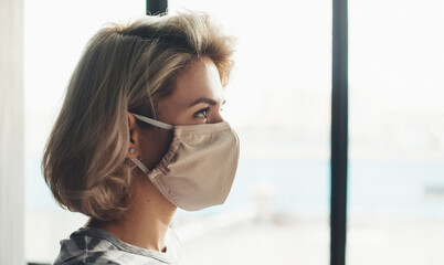 Side view photo of a blonde woman with medical mask on face sitting near the window
