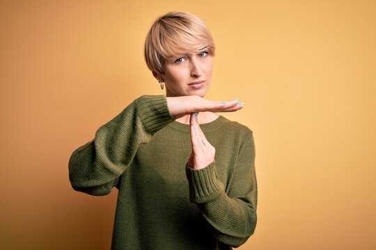 Young blonde woman with modern short hair wearing casual sweater over yellow background Doing time out gesture with hands, frustrated and serious face