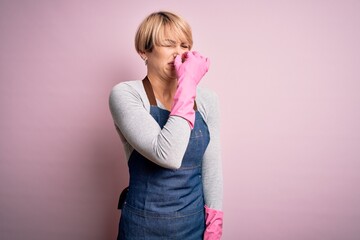 Young blonde cleaner woman with short hair wearing apron and gloves over pink background smelling...