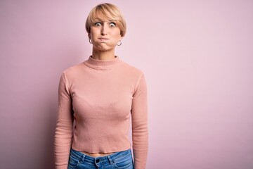 Young blonde woman with short hair wearing casual turtleneck sweater over pink background puffing cheeks with funny face. Mouth inflated with air, crazy expression.