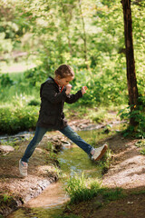 A Happy young girl jumping over a small river against the background of the forest. Childish sports. Caucasian, outdoor. Joys and games of childhood, outdoors activities concept. Selective focus.