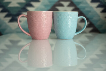 Pink and blue mugs on a white table. The table reflects objects and background. The background is bright in an ethnic style