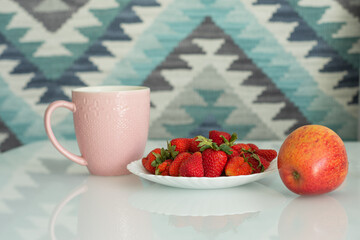 A pink Cup of tea, a white plate with strawberries and a red Apple on a white table