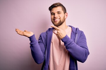 Young blond man with beard and blue eyes wearing purple sweatshirt over pink background amazed and smiling to the camera while presenting with hand and pointing with finger.