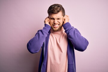Young blond man with beard and blue eyes wearing purple sweatshirt over pink background covering ears with fingers with annoyed expression for the noise of loud music. Deaf concept.