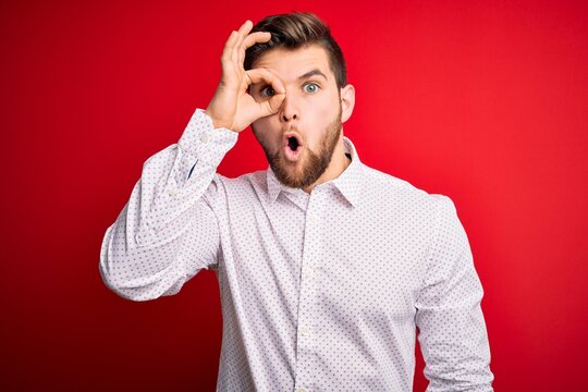 Young blond businessman with beard and blue eyes wearing elegant shirt over red background doing ok gesture shocked with surprised face, eye looking through fingers. Unbelieving expression.