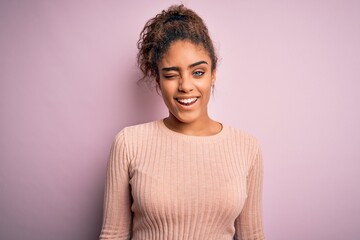 Young beautiful african american girl wearing casual sweater standing over pink background winking...