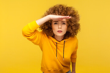 Portrait of curious curly-haired woman in urban style hoodie holding hand above eyes and peering...