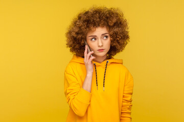 Fototapeta na wymiar Portrait of upset curly-haired woman in urban style hoodie calling friend, talking on smartphone with serious unhappy expression, discussing bad news. indoor studio shot isolated on yellow background