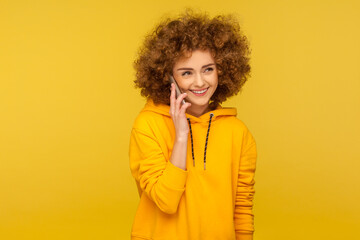 Fototapeta na wymiar Portrait of cheerful curly-haired woman in urban style hoodie calling friend, talking carefree on smartphone and smiling joyfully, discussing great news. studio shot isolated on yellow background