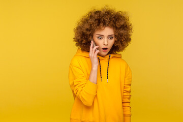 Fototapeta na wymiar Portrait of surprised curly-haired woman in urban style hoodie calling friend, talking on smartphone with amazed expression, discussing shocking news. indoor studio shot isolated on yellow background